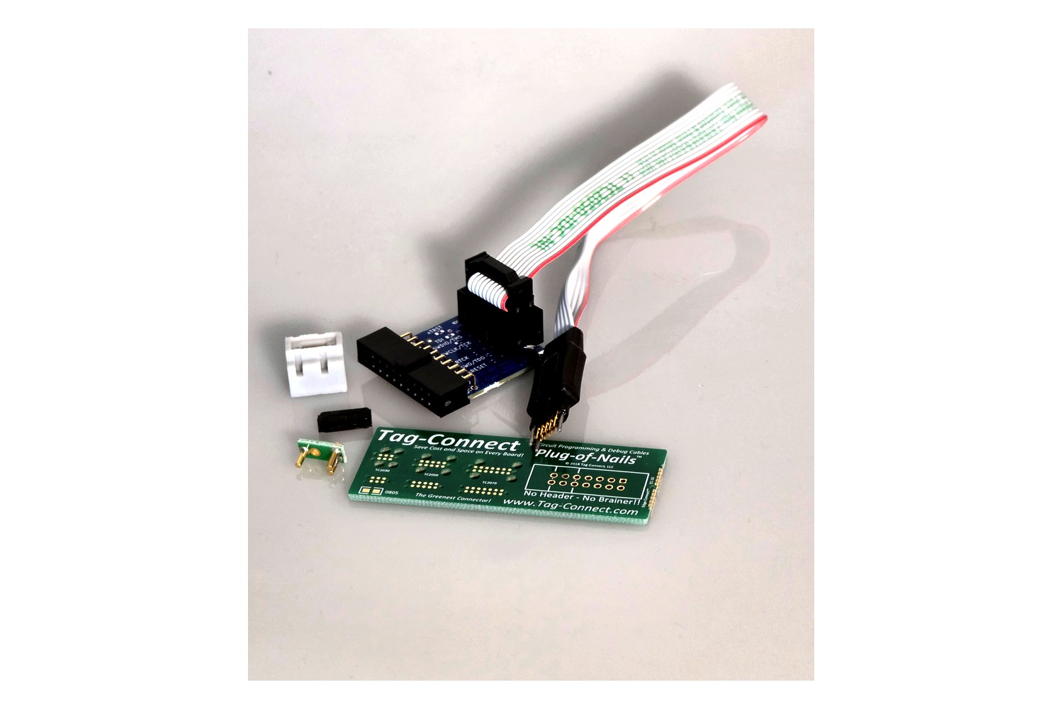 The contents of the set: the 10-pin Plug-of-Nails™ cable with ribbon lead, the blue adapter board with its 20-pin ARM connector, clip and grip as retainers, the protective cap and the green demo board with the Tag-Connect footprints.